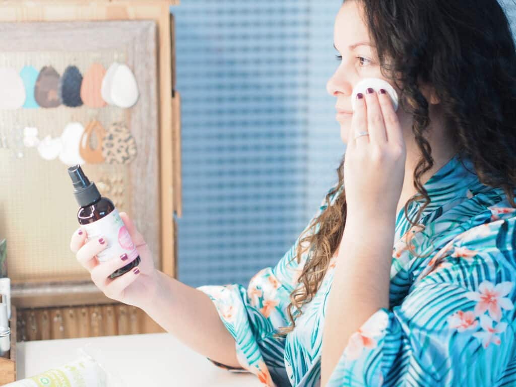 Delilah Orpi the Holistic Enchilada applying green beauty product in a glass bottle using a cotton pad at table.