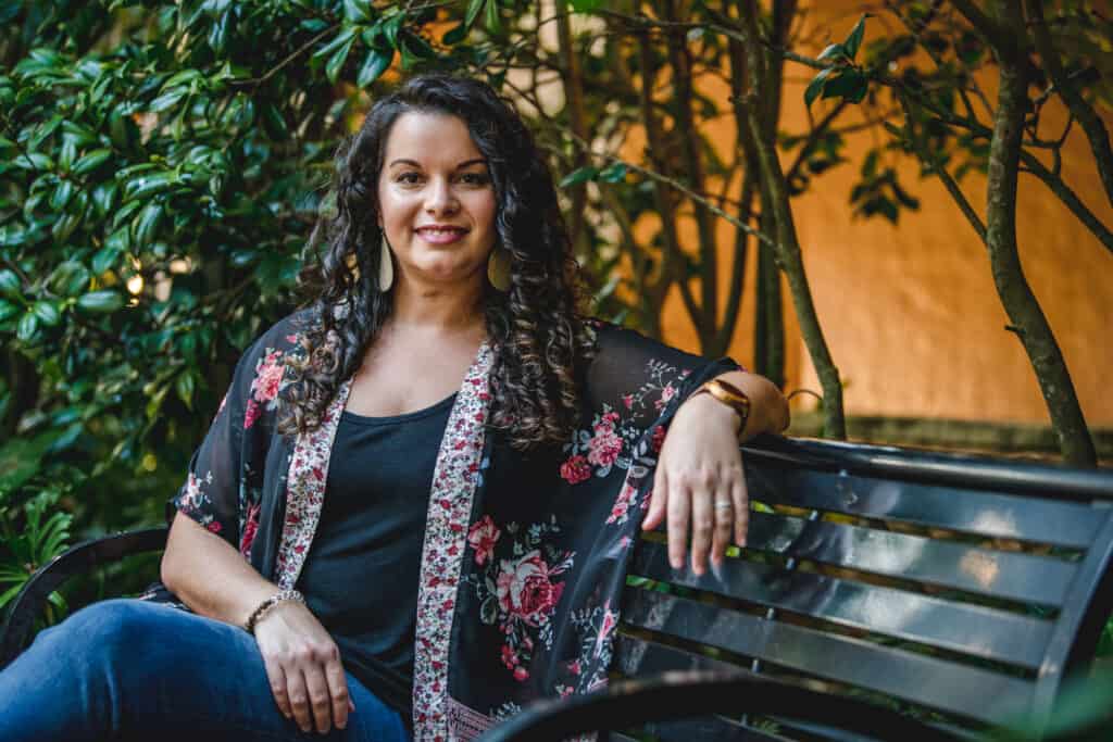 Clean beauty blogger curly hair expert Delilah Orpi sitting on bench in a garden facing the camera smiling