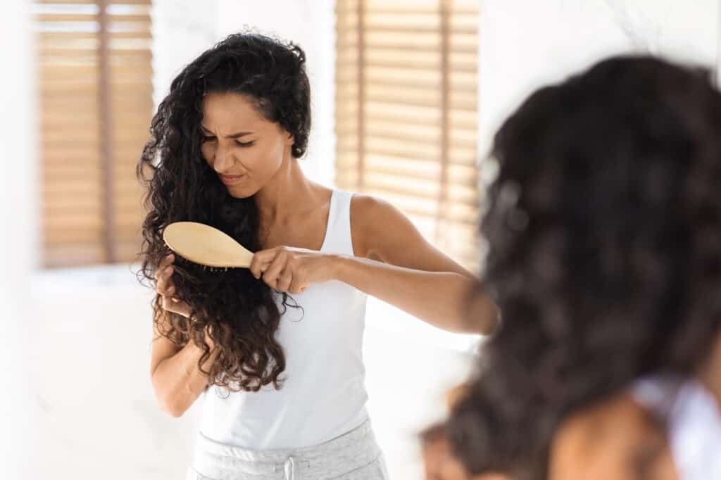 woman with matted hair trying to detangle with a brush - she is brushing her long curly hair with a brush and looks frustrated