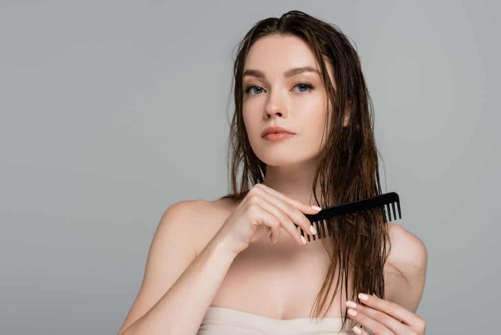 woman showing how to detangle matted hair with a comb - she is holding a black wide tooth comb and combing through her long brown hair