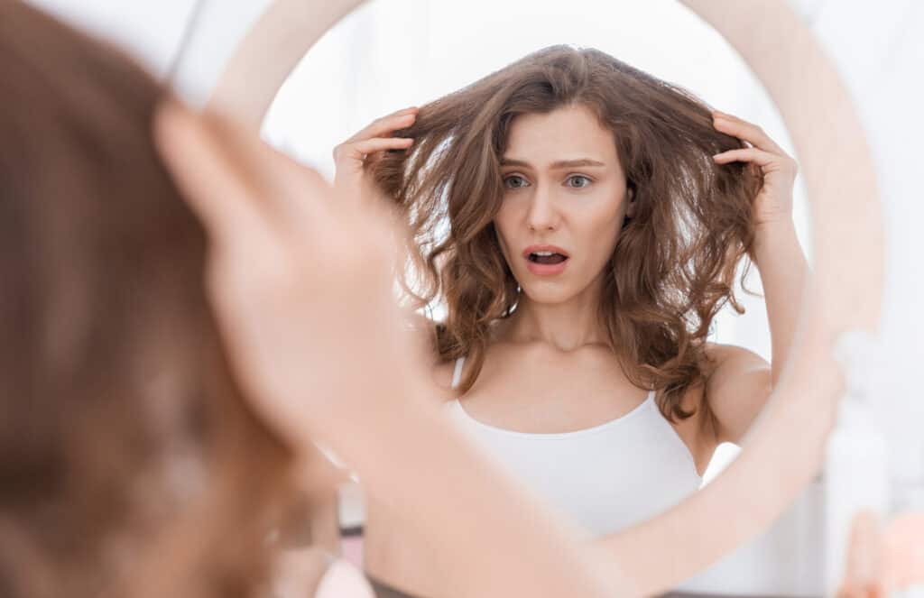 Shocked young woman holding her waxy hair, looking at mirror and asking "why does my hair feel waxy?" with buildup from polyquaternium products